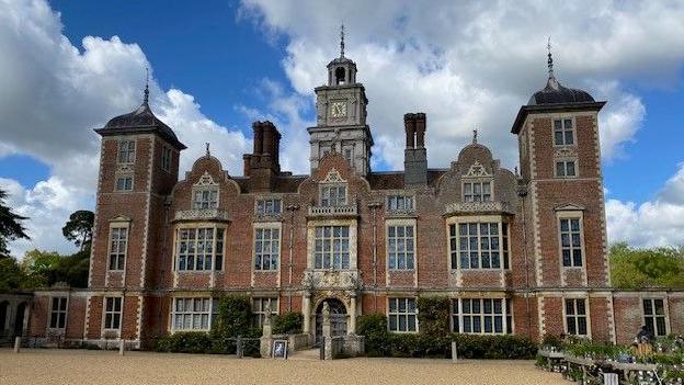 A red-brick three-storey grand hall with towers in the centre and corners. It stands in front of a gravel driveway.