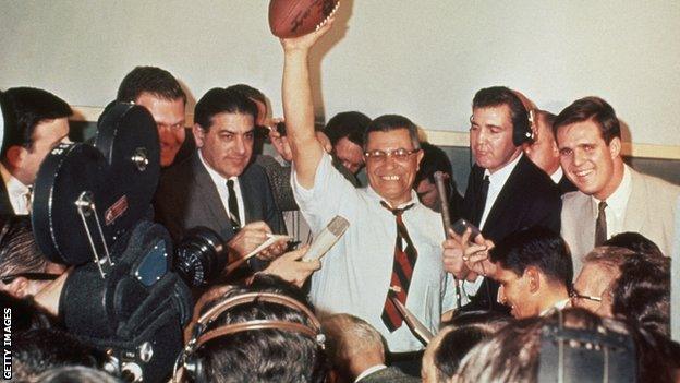 Green Bay Packers coach Vince Lombardi raises a football in victory, surrounded by reporters covering the first Super Bowl in 1967.