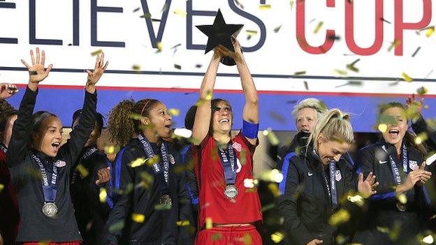 Carli Lloyd of the United States holds up the winners trophy as her team celebrates onstage after winning the SheBelieves Cup