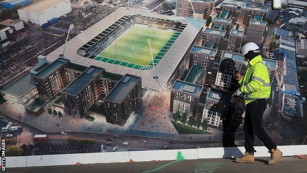 A construction worker walks past a hoarding displaying an impression of the completed new stadium at Plough Lane