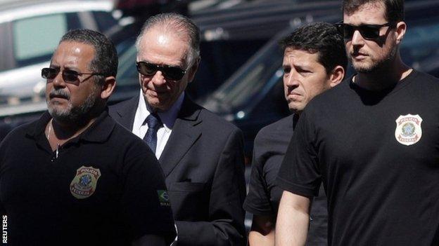 The President of the Brazilian Olympic Committee pictured between federal officers, wearing a suit and sunglasses