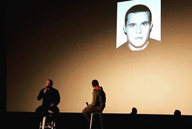 John McAvoy speaking at a public event, with his 2005 mugshot on screen behind him