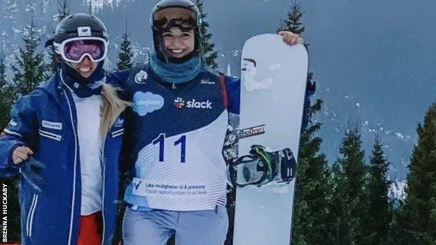 Two female para snowboarders with a snowboard standing on snow with mountains in the background