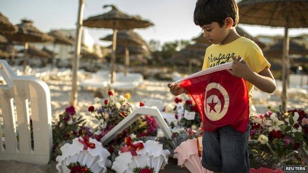 A boy holds a Tunisian flag as he stands near bouquets of flowers laid at the beachside of the Imperial Marhaba hotel