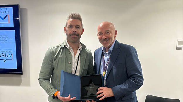 Two men stand next to each other and smile at the camera. The man on the left has light spiky hair and wears a green jacket and a white shirt. The man next to him is bald and has a blue shirt and suit jacket. They both hold a box with a star-shaped award in it.
