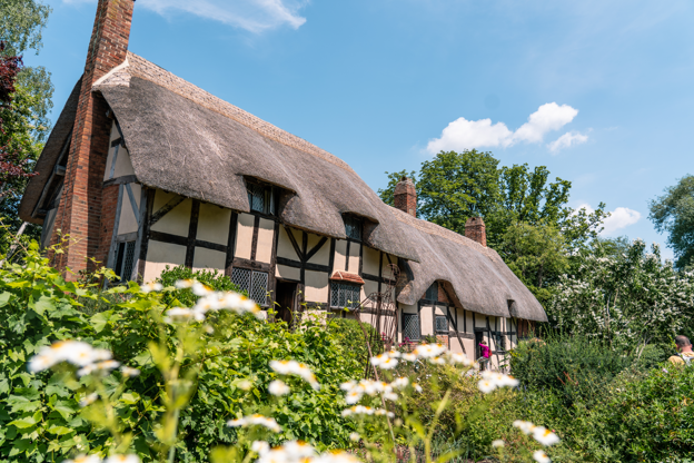 A thatched 15th-century house surrounded by flowers and trees. The walls are white with brown beams.