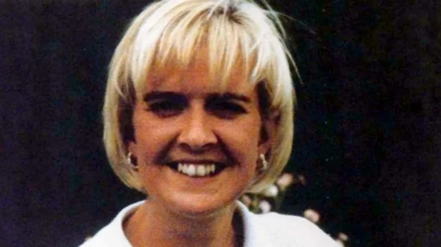 The picture shows a head shot of a smiling woman with a blond bob, wearing a white shirt with earrings.