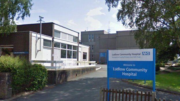 A blue and white sign that reads "Ludlow Community Hospital". In the background are large grey, brown and white buildings, with a large tree on the right hand side.