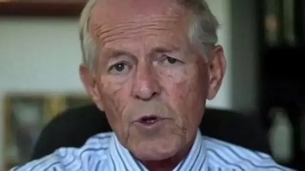 A screenshot of John Smyth being interviewed. He is wearing a stripey white and blue shirt and in a chair, which is blurred because it is in the background and not in focus.