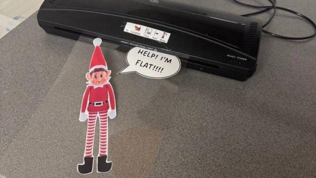 The elf wears a red hat with a white trim and bobble, a red coat with a white collar and gloves and a black belt with red and white striped legs and black boots. It is flattened and laminated in a clear plastic. There is a speech bubble coming from its mouth saying "help! I'm flat"