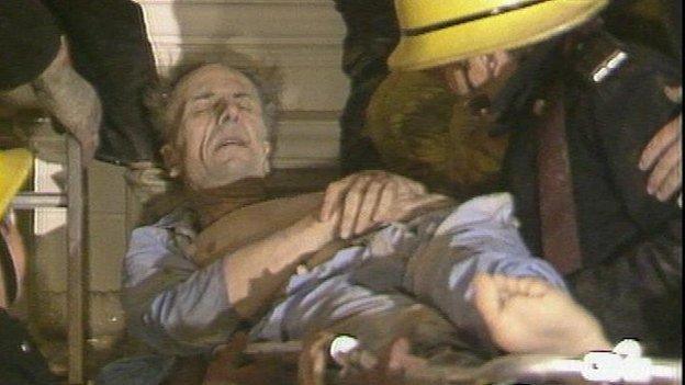 Norman Tebbit, with his eyes closed and wearing blue pyjamas, is stretchered out of the Brighton bomb hotel. He has short, balding dark hair and appears to be grey with dust. Firefighters in yellow helmets surround him as they carry him.