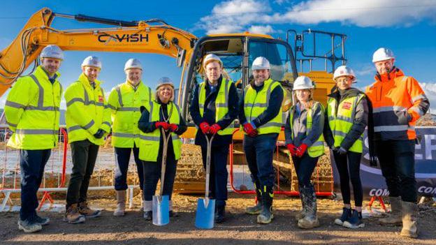 A line of nine people in mostly yellow hi-vis jackets. They all have white hard hats.  A man and a woman in the centre of the line-up have blue spades. They are standing on soil. There is a yellow digger behind them.