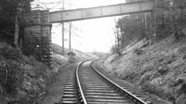 A black-and-white image of a railway line with a narrow metal bridge crossing it. 