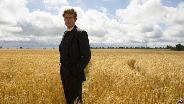 James Norton dressed as an Anglican clergyman in the 1950s and standing in a golden corn field. He is wearing a black suit, with a white dog collar and looking towards the viewer. His hands are in his pockets. Beyond the flat field, it is edged with trees and above is a cloud covered blue sky.