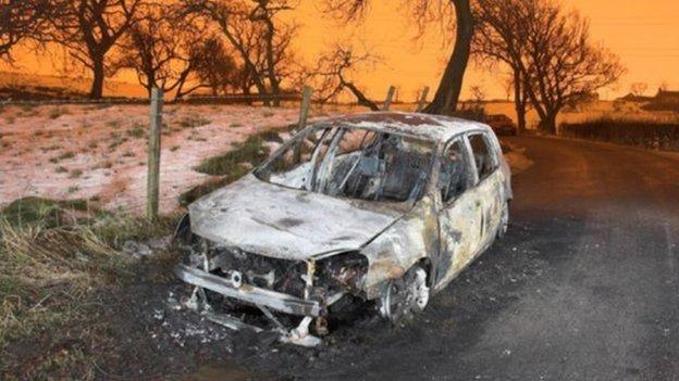 The charred, grey remains of the Volkswagen Golf used in the murder of Kevin Carroll. The vehicle, which has been reduced to a burnt out shell, is photographed at the side of a country road. Snow is visible in the fields,  behind a wire fence. The sky in the background is a striking red/orange.  