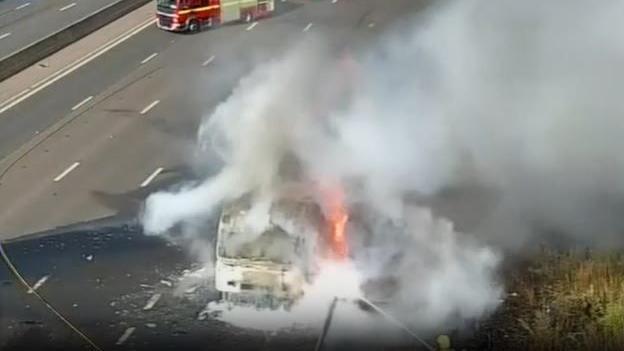 A lorry heavily on fire on a motorway with grey smoke surrounding the vehicle