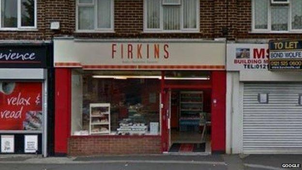 A shop front in a mid-20th Century parade of shops with Firkins written in red on a white sign above the door.
 
