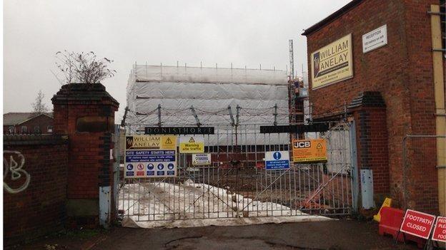 Locked steel-bar gates at a factory with a building behind covered in scaffolding, and plastic and site safety notices placed on the gates.  