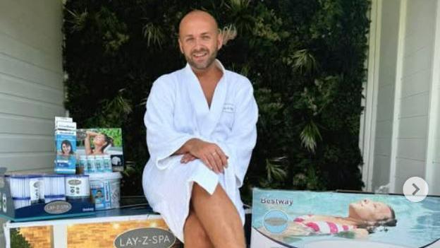 A man sits outside, surrounded by boxes containing the equipment needed to set up a hot tub. He is wearing a white dressing down. He has a shaved head and stubble.