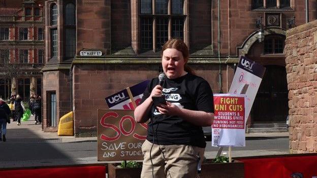 A woman in a black T-shirt with a white graphic on the front and brown trousers is talking into a microphone while looking at her mobile phone. Behind her are placards about university cuts