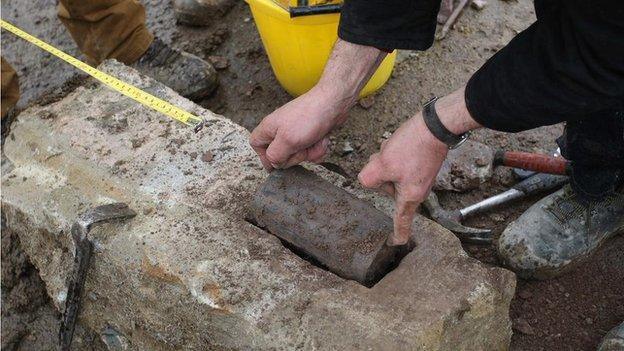 a lead capsule is removed from rock by archaeologists. a pair of hands is seen lifting it the capsule out of the rock. a tape measure is seen in the background, as are a workers boots.