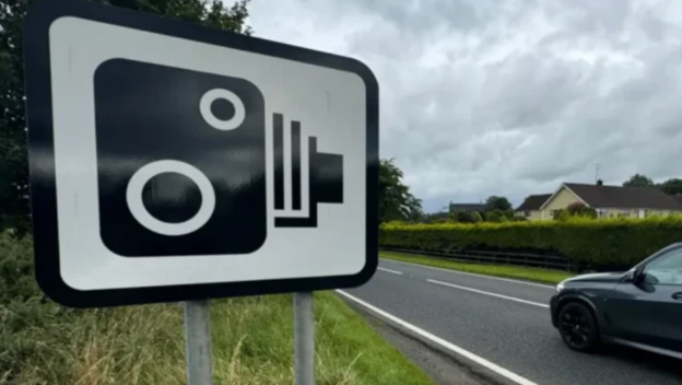 A stock image of a speed camera sign beside a B road. A car can be seen driving past the sign.