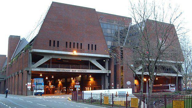 Brick building with ground floor showing two vast openings with grey frames and orange lights within. A bus is visible in each opening. The legend "Greyfriars Bus Station" is visible over the opening on the left. There is a triangular section above in which windows are visible and the side to the left is made of glass. 