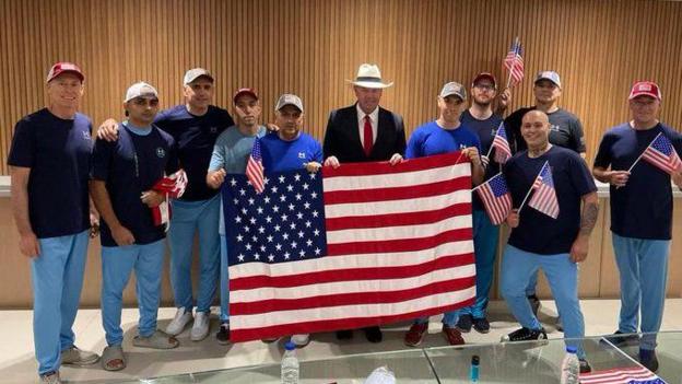 Several men in jeans, blue t-shirts and caps hold up a large American flag and pose for photo