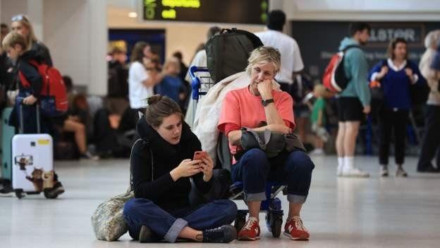 Two women wait at an airport terminal. One is squatting on the floor looking at her phone. The other is sitting on a luggage trolley with her head resting on her hand.