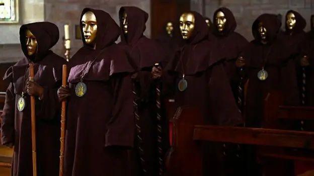 The image shows a procession of cloaked traitors wearing gold masks