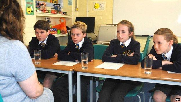 4 children sit at desks looking at an adult facing them