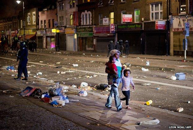 A woman walks through the debris with two children as riot police try to contain a large group of people on a main road in Tottenham, north London 6 August 2011