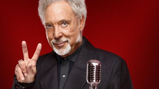 Tom Jones appears in a publicity photo for BBC The Voice. He is wearing a smart black suit and is stood behind a recording studio microphone. He is making a peace sign with his fingers, representing the letter V in The Voice.