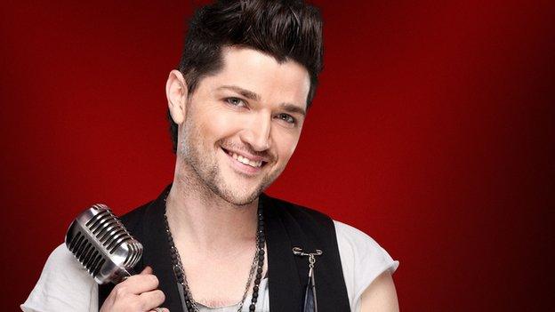 Danny O'Donoghue in his publicity photograph for BBC The Voice. He is wearing a white t-shirt and a smart black waistcoat. His hair is coiffed into an impressive quiff! He is holding a retro microphone and smiling.