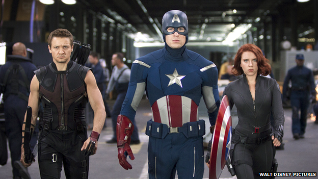 Three of the Avengers march through a futuristic tunnel in what looks like a secret government base. The man in the centre is dressed as a superhero in red, blue and white. A man and woman dressed in black stand either side of him.