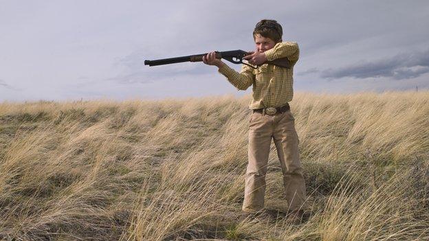 A young boy in a field aims a rifle