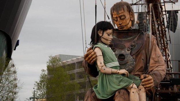 A giant marionette of a girl sits in the lap of a giant diver