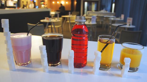 Popular soft drinks lined up with the amount of sugar they contain stacked next to them in sugar cubes