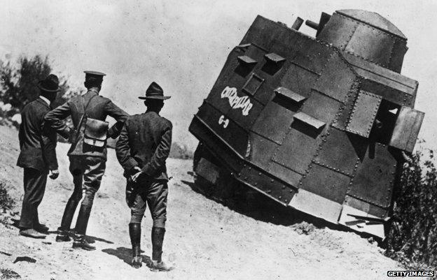 Three men look on as a US tank appears to be stuck on the side of a road