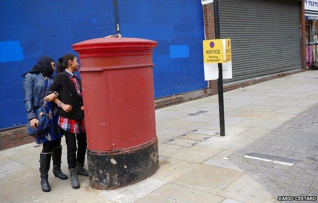 Two women hide behind a postbox