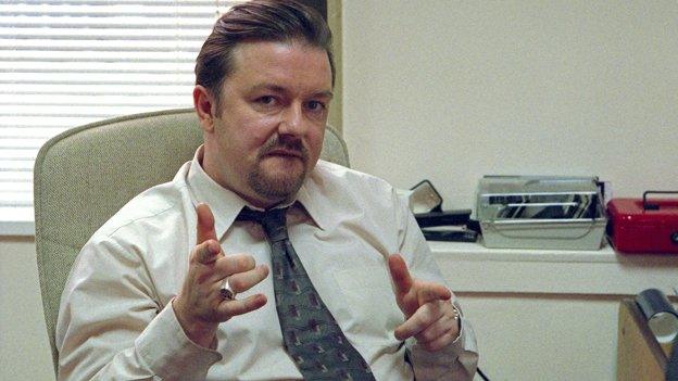 Ricky Gervais as David Brent in The Office, sat behind his desk pointing at the camera with both hands