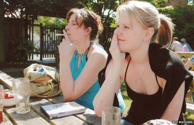 Two women are sat in a beer garden, one is smoking a cigarette