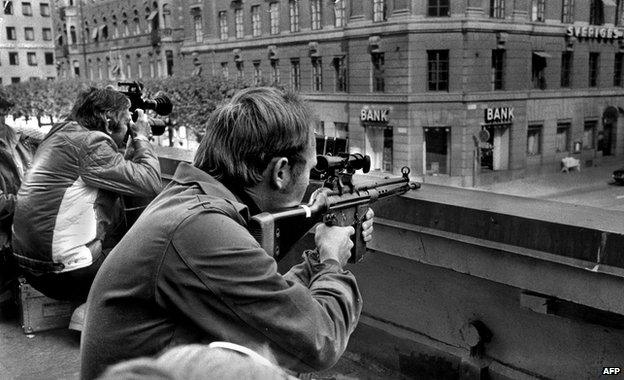 Two policemen hold guns, poised opposite a Kreditbanken building