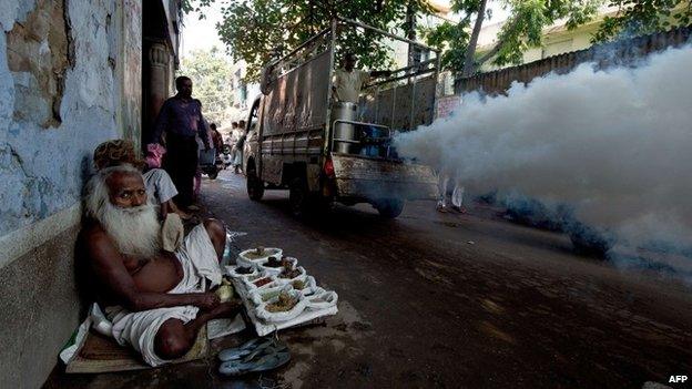 An elderly Indian man sells Ayurvedic medicines at the roadside as a municipal corporation worker fumigates the area to prevent mosquitoes from breeding in New Delhi on September 27, 2013.