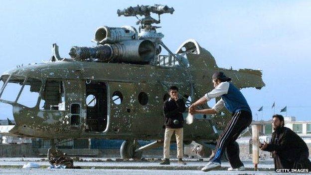 Afghan cricketers take part in a match on a patch of ground in front of a destroyed helicopter