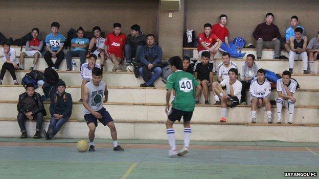 Two men playing football in a gym, with others watching