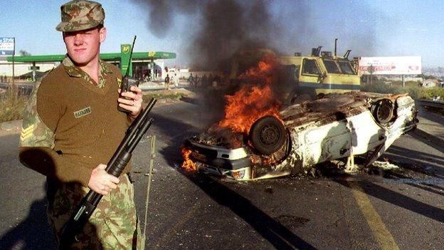 A South African policeman stands near a burning car outside Soweto (19 April 1993)