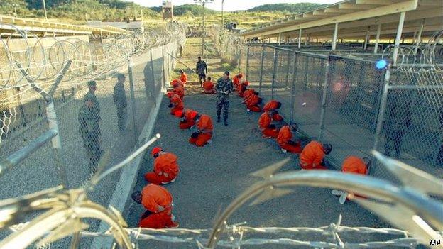 Detainees in orange jumpsuits sit in a holding area under the watchful eyes of military police at Camp X-Ray at Naval Base Guantanamo Bay, Cuba, during in-processing to the temporary detention facility on January 11, 2002