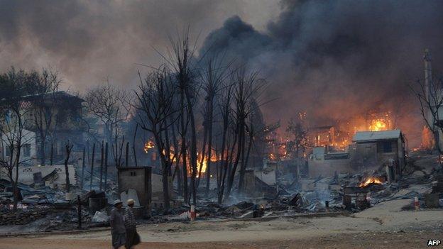 Residents walk past buildings burning in riot-hit Meiktila, central Burma, 21 March 2013