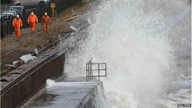 A wave crashes onto a damaged section of railway track, as repair workers walk nearby